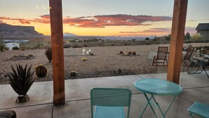 A patio with a view of a desert landscape at sunset, featuring a concrete floor, metal chairs, and a small table.