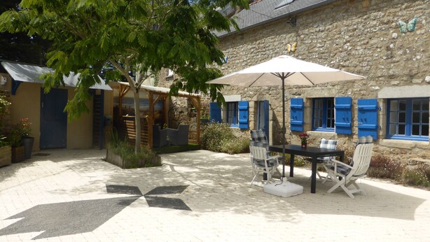 A stone building with a patio area featuring a table and chairs under a canopy, surrounded by potted plants and a tree.
