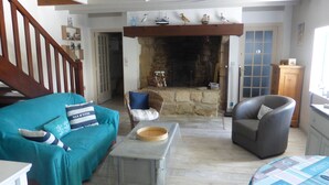 A living room with a stone fireplace, a wooden staircase, a blue sofa, a grey armchair, and a small table with a bowl.