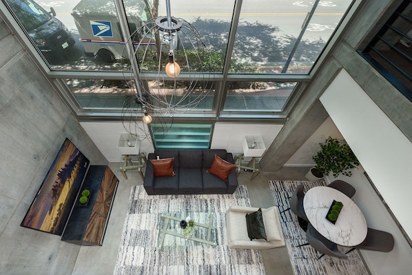 A modern living area with a sofa, coffee table, and dining table, a large window with a view of the street, and a unique light fixture.