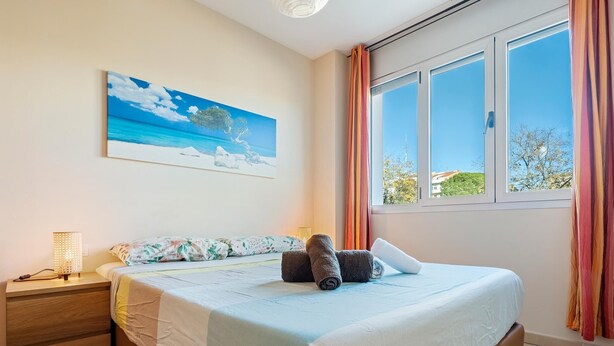 A bedroom with a bed, a nightstand, a lamp, a painting of a beach, and a window with curtains. - Apartment with terrace & pool 5min beach (Sant Feliu de Guíxols)