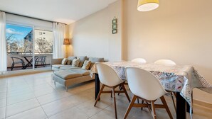 A modern living room with a sofa, dining table, and chairs. There is a balcony with a view of buildings and trees. - Apartment with terrace & pool 5min beach (Sant Feliu de Guíxols)