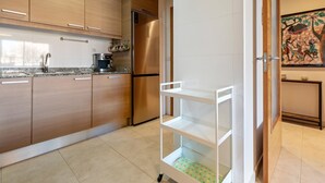 A modern kitchen with wooden cabinets, a stainless steel refrigerator, a coffee maker, and a white shelving unit. - Apartment with terrace & pool 5min beach (Sant Feliu de Guíxols)