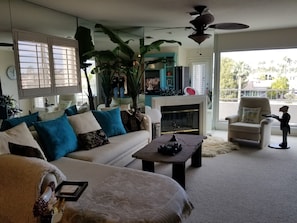 A living room with a sofa, coffee table, and a fireplace. There is a large plant, a ceiling fan, and a view of the outdoors.