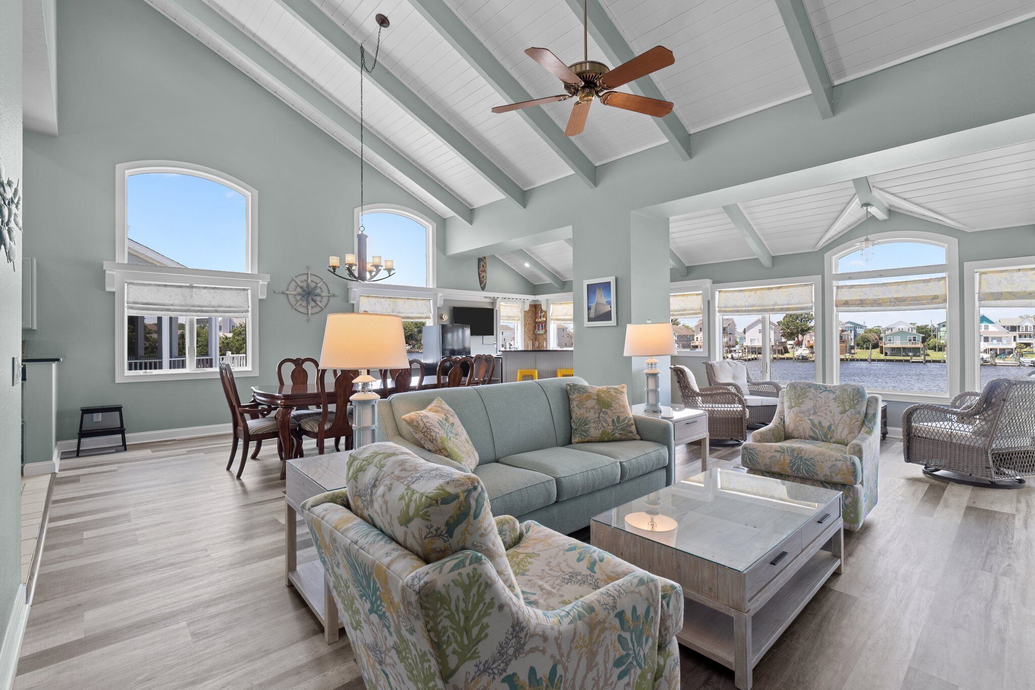 A spacious living room with a large sofa, two armchairs, a coffee table, and a dining area with a table and chairs. There is a ceiling fan, large windows, and a view of the water outside.