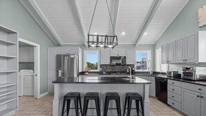Una cocina moderna con armarios blancos, una isla central con taburetes, una lámpara colgante y vistas al exterior.