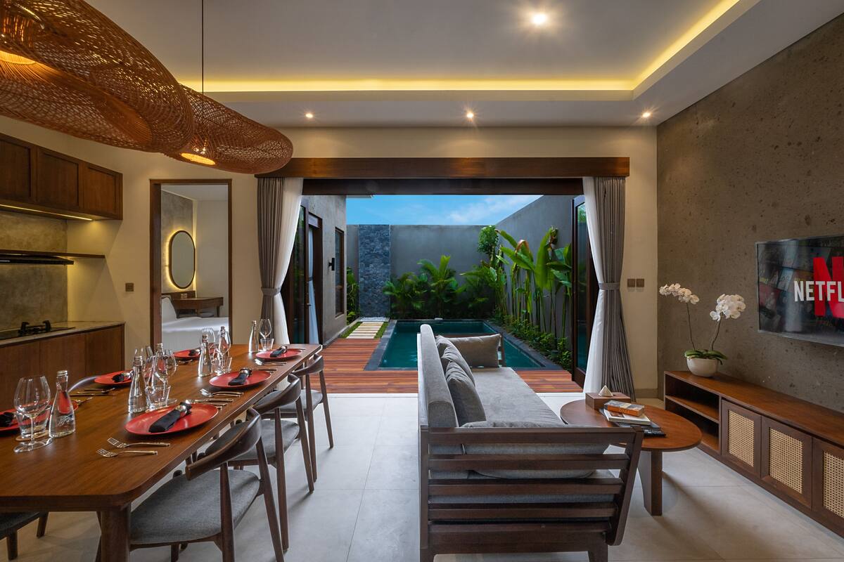 A modern dining area with a pool view, wooden furniture, and a TV on the wall.