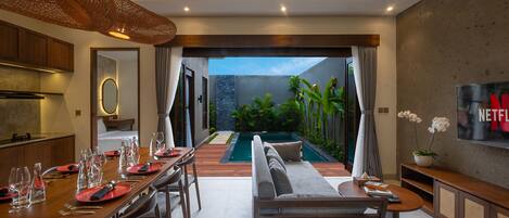 A modern dining area with a pool view, wooden furniture, and a TV on the wall.