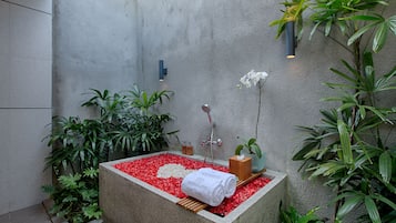 A spa area with a stone bathtub surrounded by red pebbles, a wooden bench, and various plants.