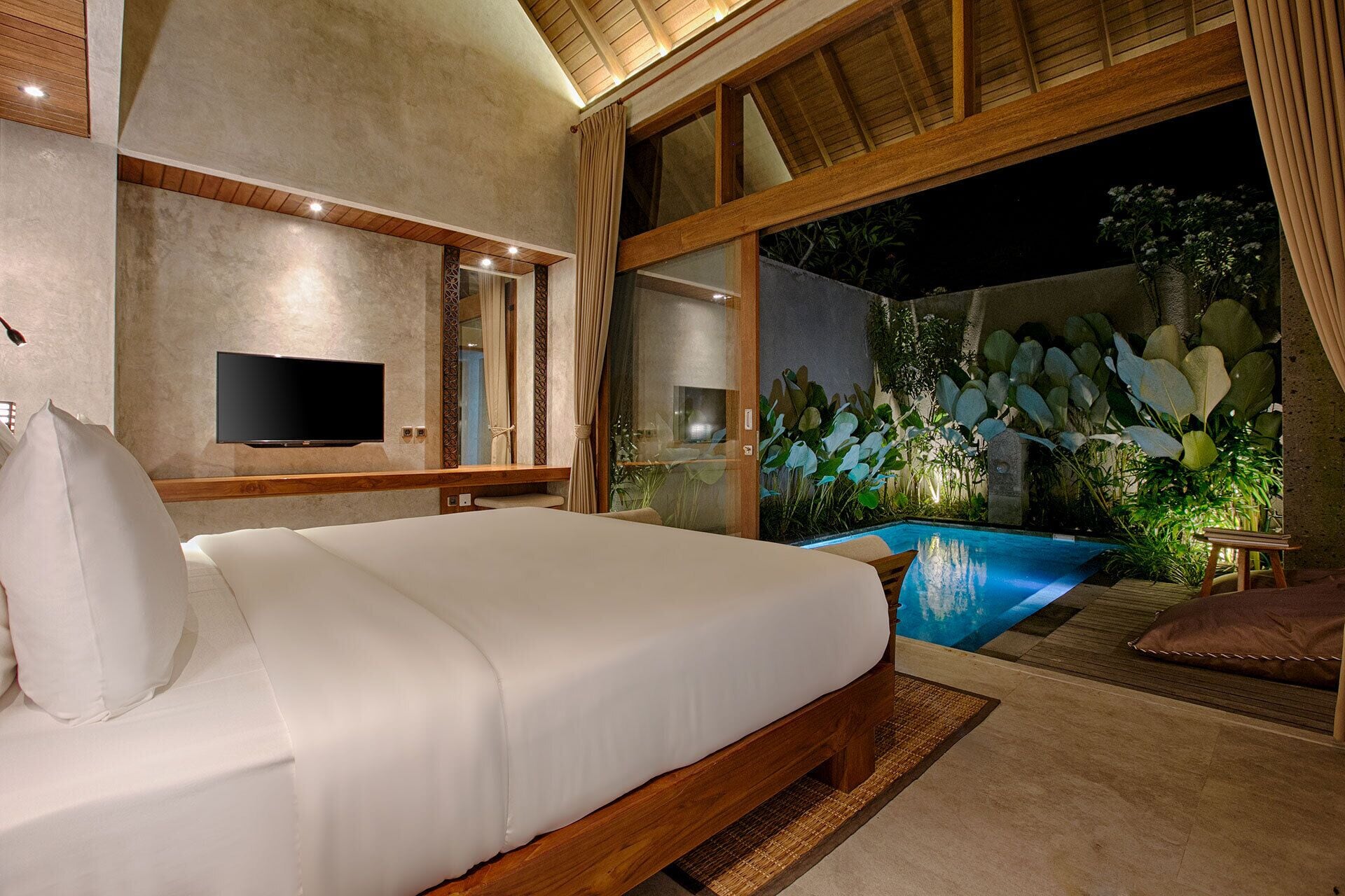 A bedroom with a bed, a TV, a window with curtains, and a view of a pool and plants.