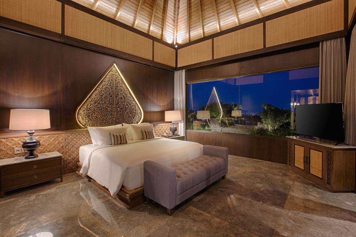 A modern bedroom with a large bed, a nightstand, a TV, and a view of a cityscape at night.