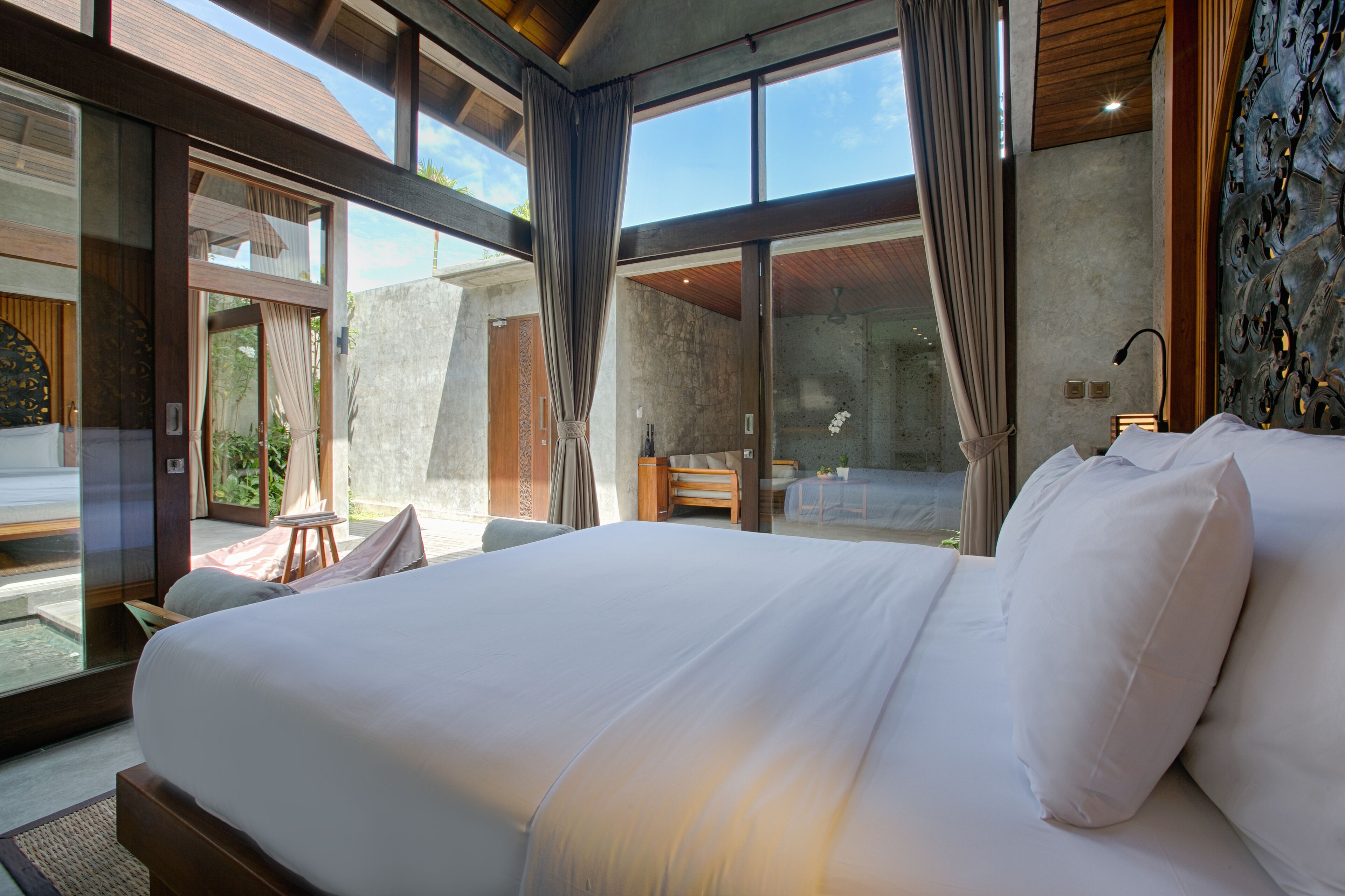 A modern hotel room with a large bed, a wooden headboard, and large windows offering a view of the outdoors.