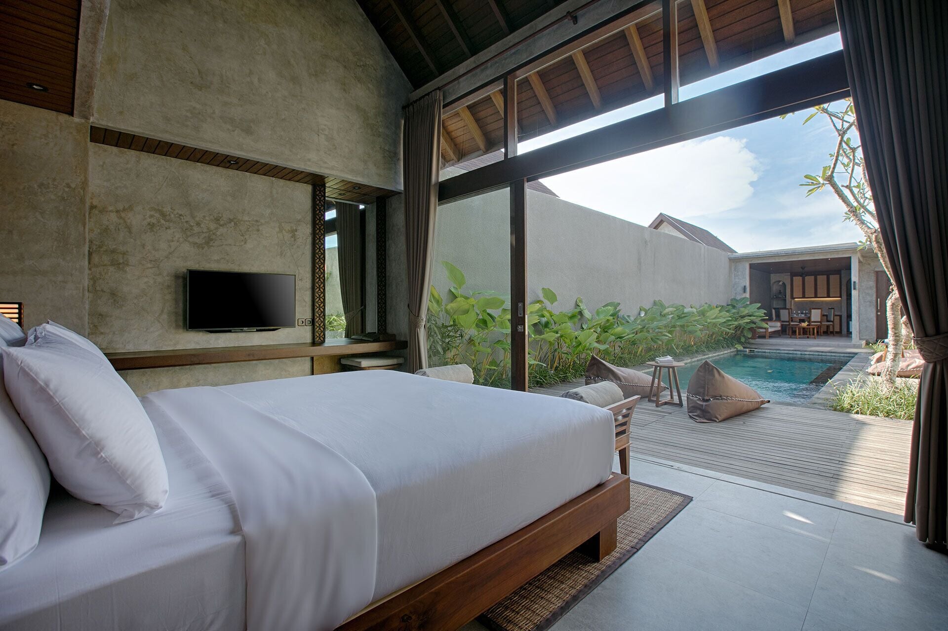 A bedroom with a large bed, a TV mounted on the wall, and a view of a pool and outdoor seating area.