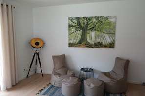 A room with a tripod floor lamp, two bean bag chairs, a small round table with two glasses, and a large forest-themed painting on the wall.