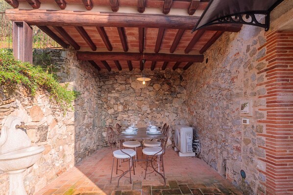 A stone-walled outdoor dining area with a wooden ceiling, a table set for four, and a white appliance nearby.
