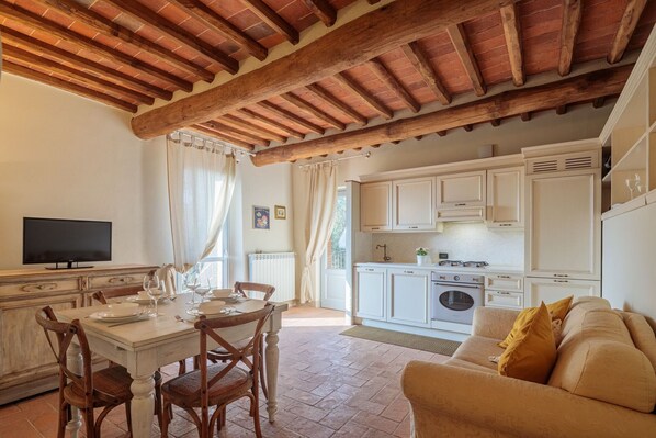 A cozy living area with a dining table, wooden chairs, a sofa, and a kitchen with white cabinets and a tiled backsplash.