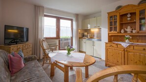 Un salon confortable avec un canapé, un téléviseur, une table à manger avec des chaises, un buffet en bois avec des portes en verre et un coin cuisine avec des placards et une cuisinière.