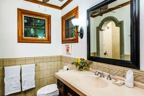 Une salle de bain avec un grand miroir, deux fenêtres, un lavabo agrémenté d’un vase de fleurs et des toilettes.