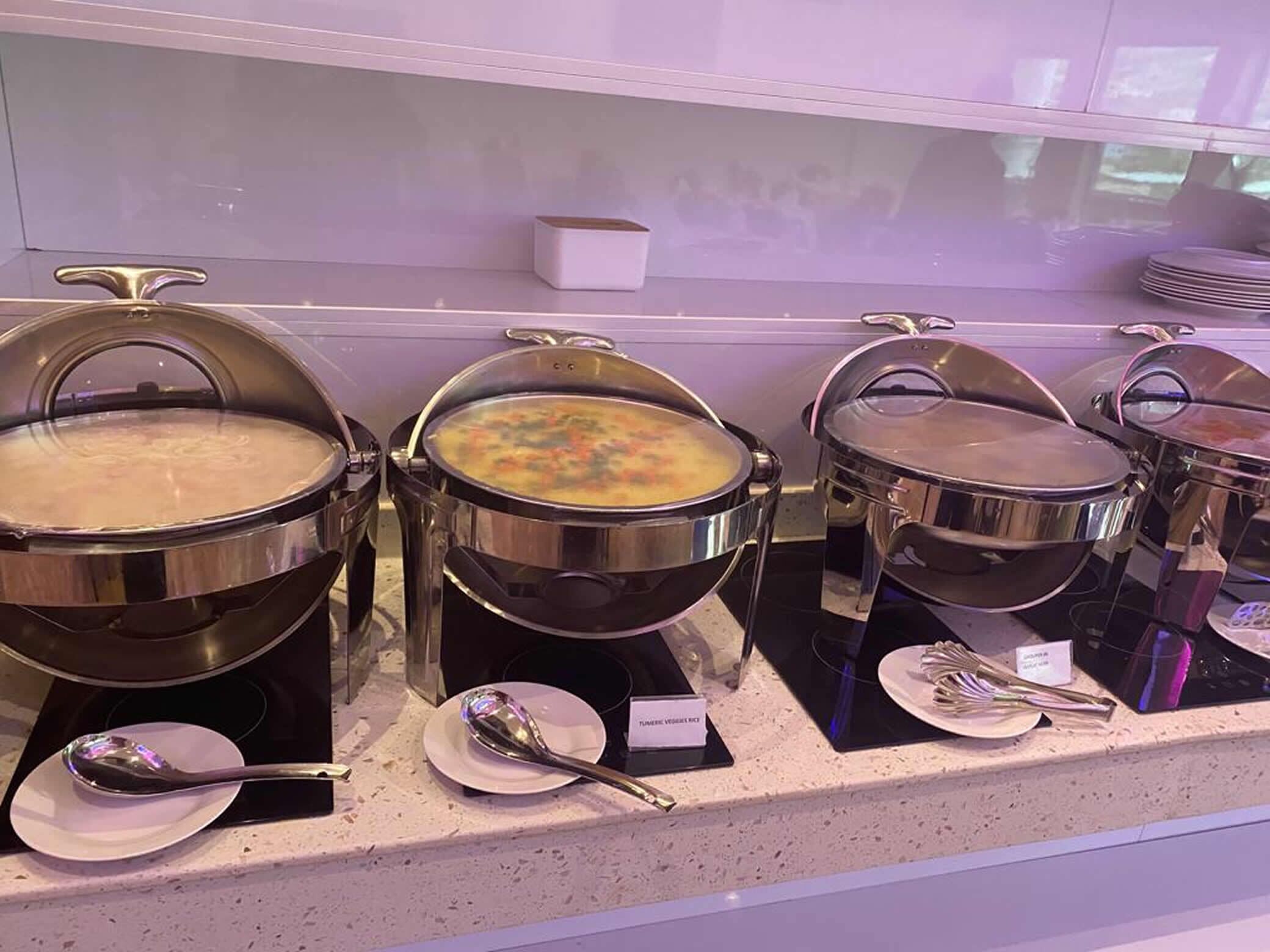 Un buffet proposant divers plats présentés dans des plats de service en acier inoxydable, chacun accompagné d’une cuillère et d’une étiquette indiquant le nom du plat.
