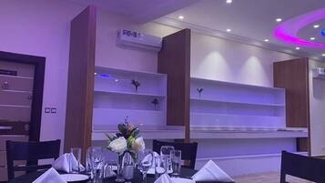 A dining area with a round table set for four, surrounded by chairs. There are air conditioning units on the walls, and the ceiling features recessed lighting with a purple hue.