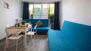 Ein Zimmer mit einem blauen Sofa, einem kleinen Tisch mit Stühlen, einem Fenster mit Vorhängen und einer Heizung.