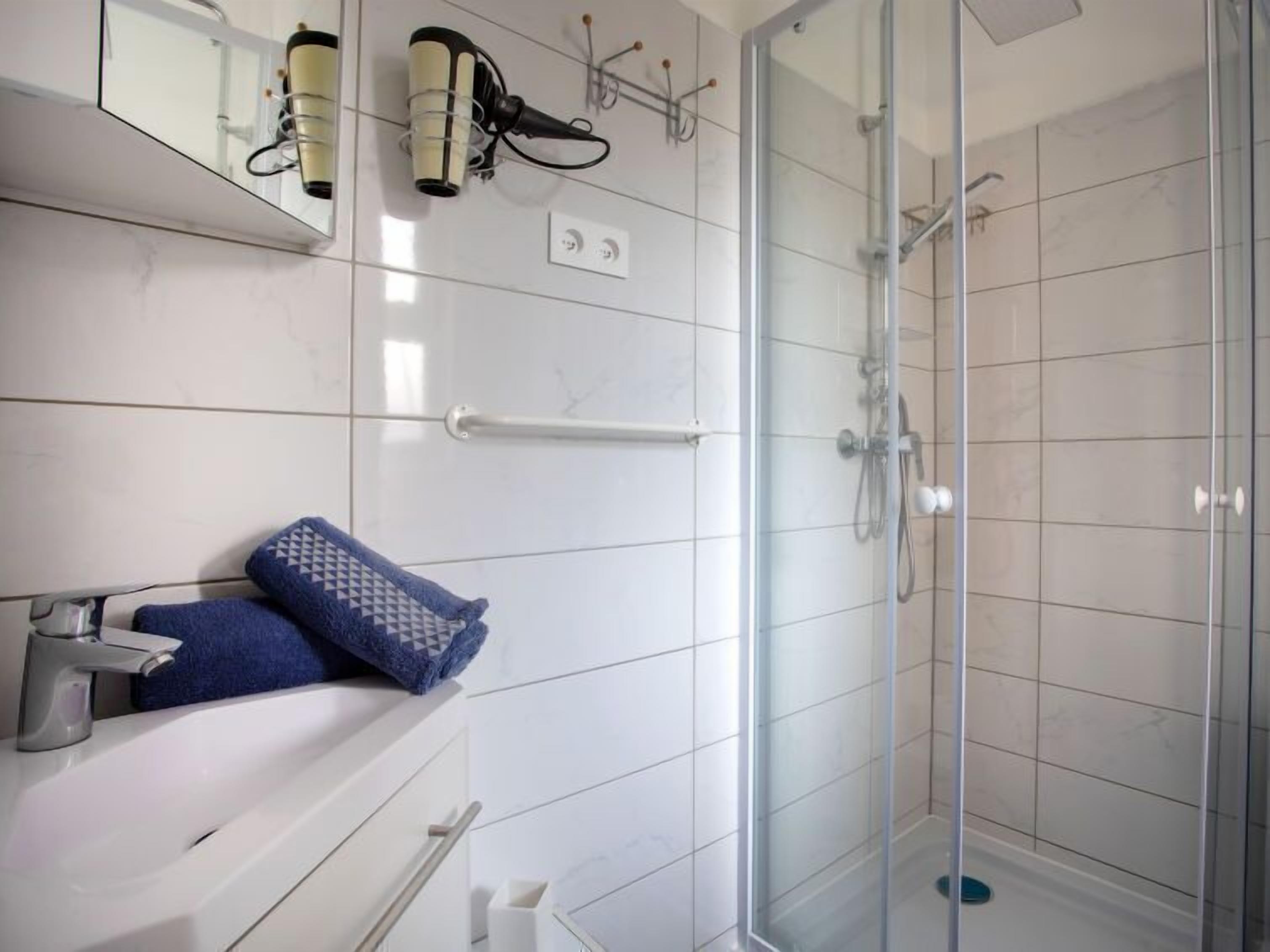 Une salle de bain moderne avec une douche à parois de verre, des murs recouverts de carreaux blancs et un lavabo blanc avec une serviette.