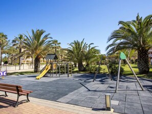 Un parque infantil con tobogán y estructuras para escalar, rodeado de palmeras y un banco.