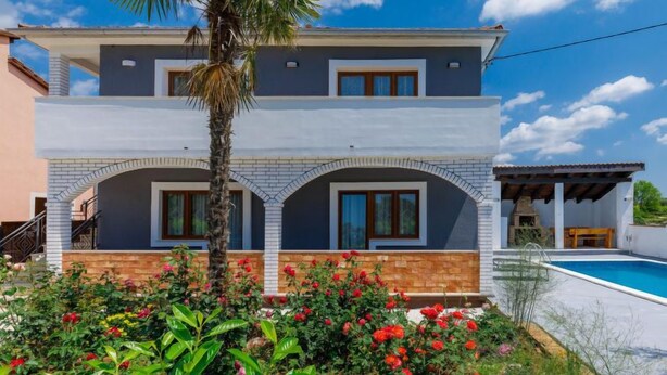 Una casa de dos pisos con balcón, piscina y jardín con flores rojas.