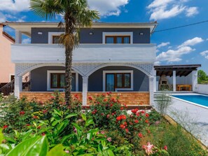 A two-story house with a balcony, a swimming pool, and a garden with red flowers.