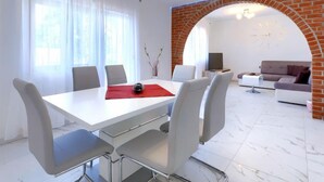 Una sala de estar moderna con una mesa de comedor blanca, sillas grises y un mantel individual rojo. Se observa una chimenea de ladrillo, un sofá blanco y un televisor al fondo.