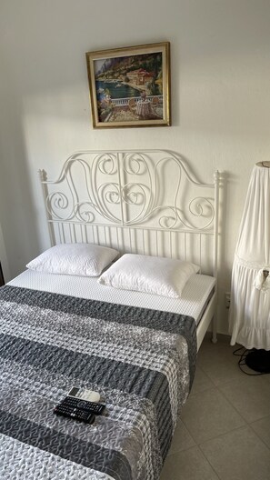 A neatly made bed with a patterned bedspread and pillows, a white metal headboard, a painting above the bed, and a white curtain on the right.