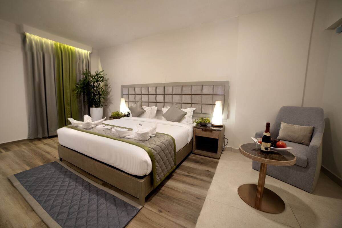 A modern hotel room with a large bed, bedside tables, a small round table with a bottle, and a grey armchair.
