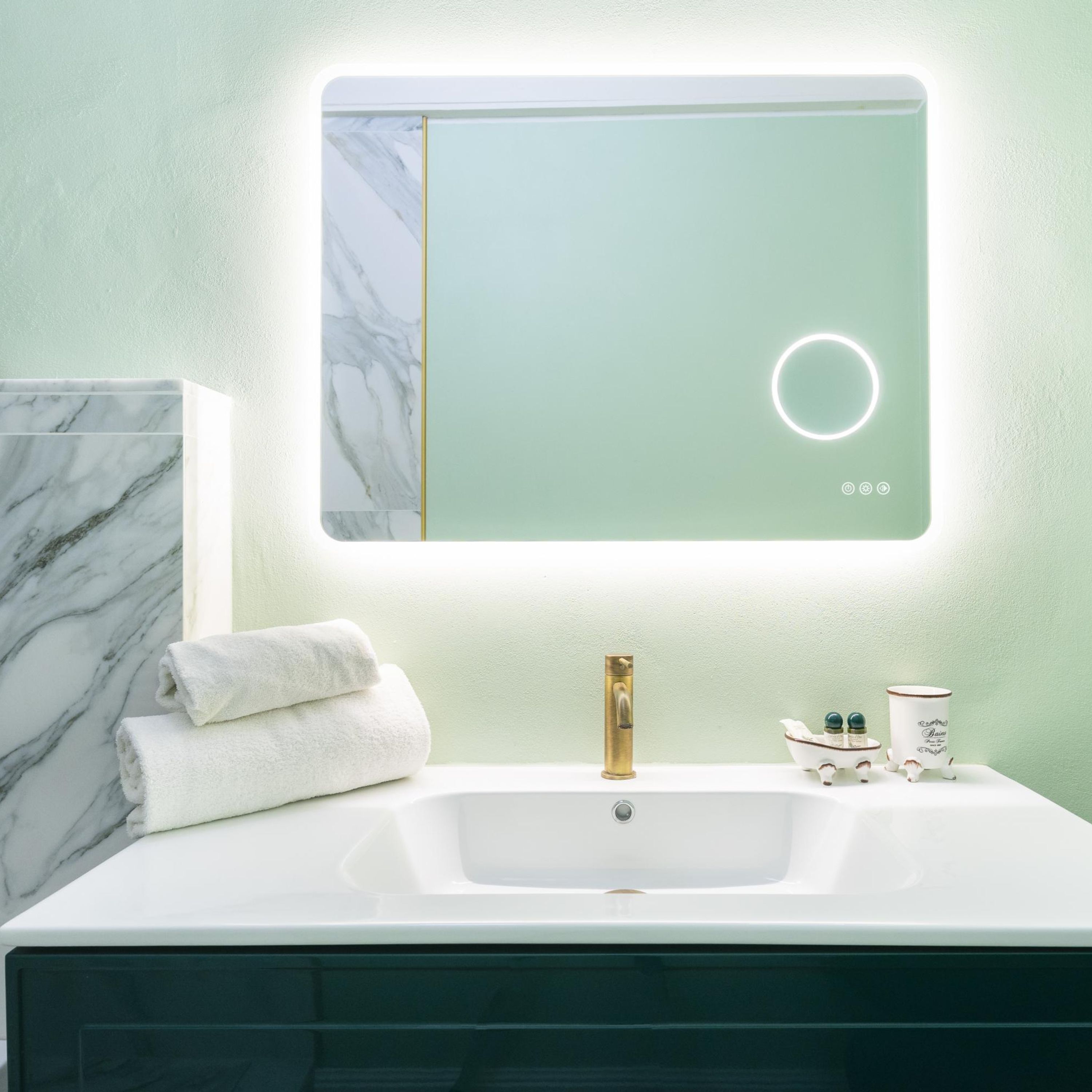 A bathroom with a marble-tiled wall, a white sink with a gold faucet, and a mirror with a circular light.