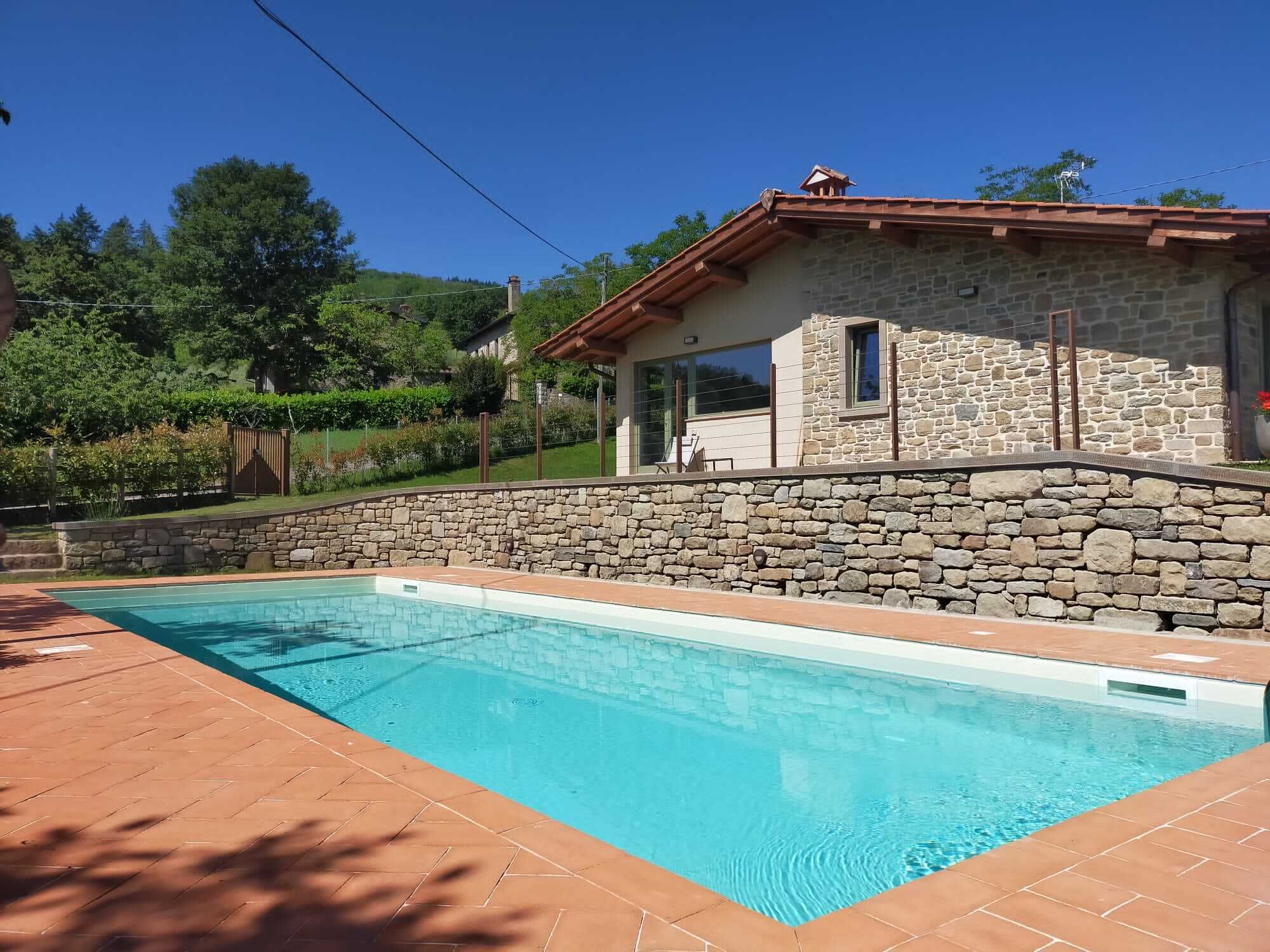 Una piscina con paredes de piedra, una casa de piedra con techo rojo y vistas a colinas verdes.