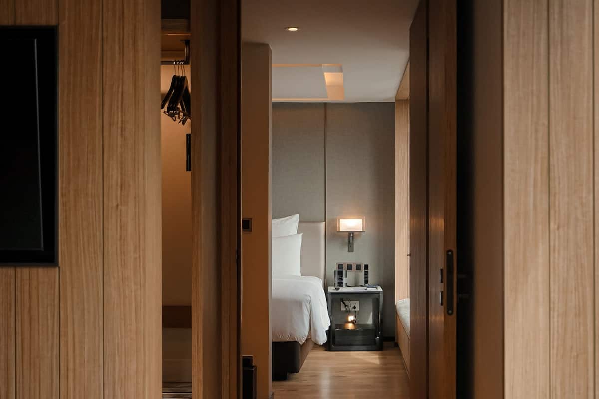 Un couloir d’hôtel aux murs en bois, un lit avec une literie blanche, une table de chevet avec une lampe et une petite commode avec une horloge.