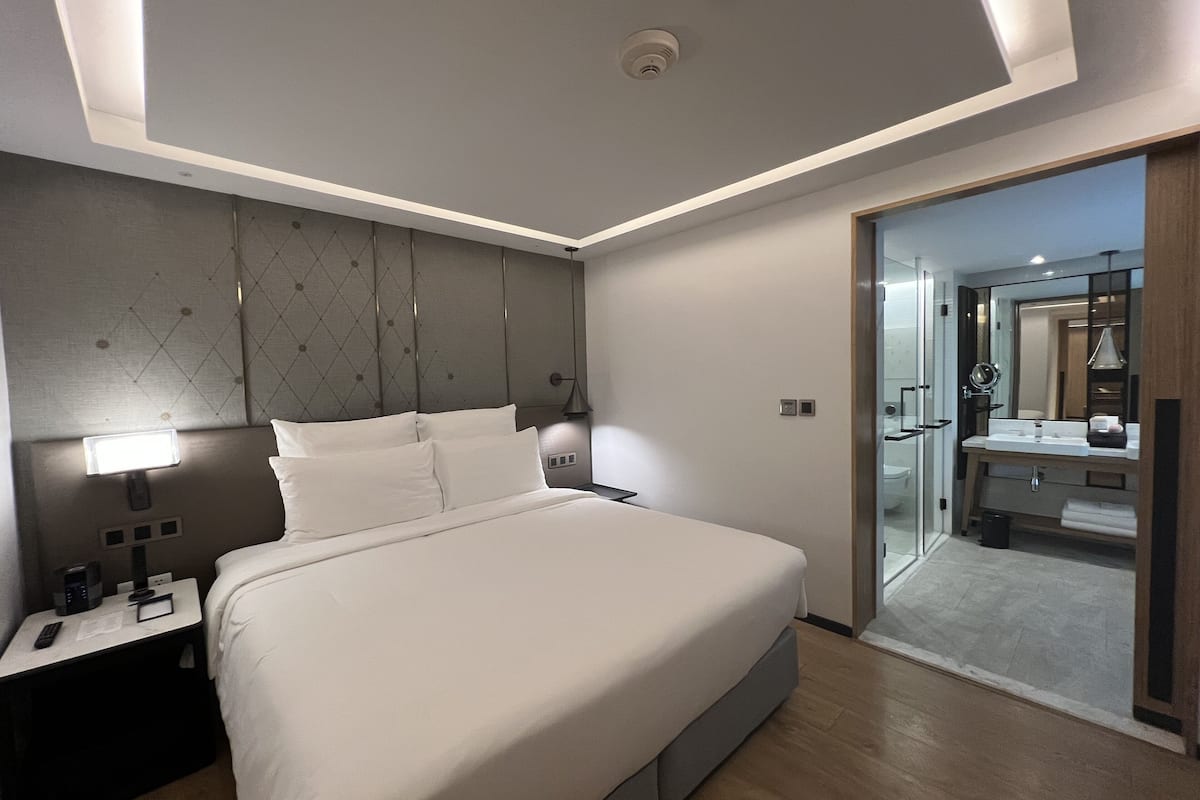 Une chambre d’hôtel moderne avec un grand lit, une table de chevet avec une lampe, une commode et une porte vitrée donnant sur une salle de bain équipée d’un lavabo et d’un miroir.