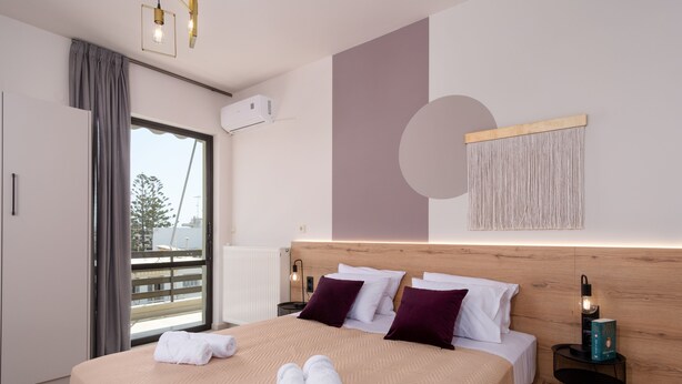 Une chambre d’hôtel moderne avec un grand lit, une tête de lit en bois, un climatiseur fixé au mur et une vue dégagée sur l’extérieur grâce à une grande fenêtre.