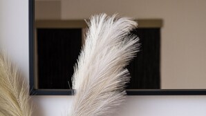 Un vase en verre transparent contenant des plumes d’herbe des pampas, posé sur une étagère en bois devant un mur uni, avec un miroir à cadre noir.