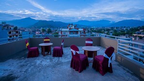 A rooftop terrace with tables and chairs, offering a view of a cityscape and mountains in the background. - Hotel Pauwa Pokhara (Pokhara)