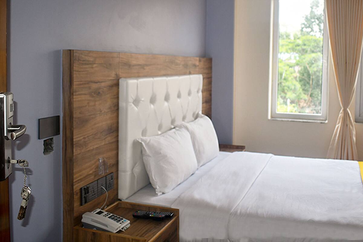 Habitació d'hotel amb un panell de capçalera de fusta, un llit amb roba de llit blanca, una tauleta de nit amb un telèfon i un comandament a distància, i una finestra amb cortines.