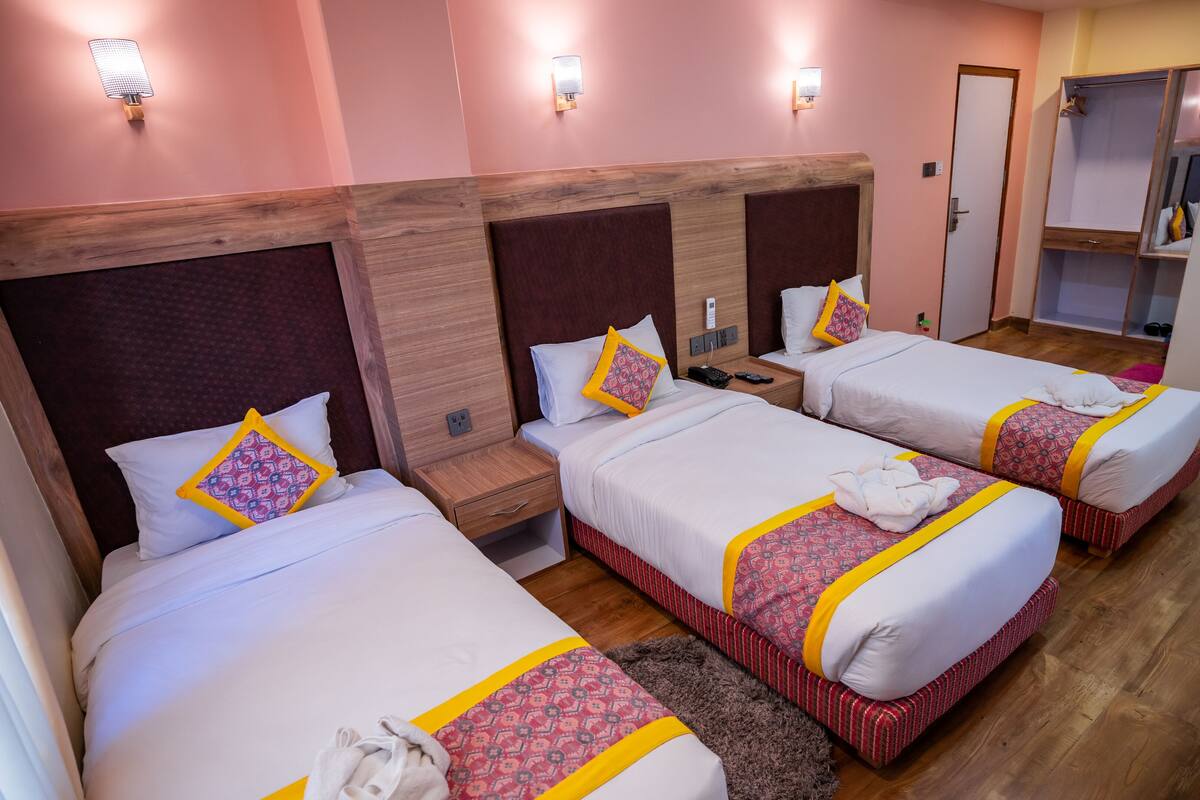 Una habitació d'hotel amb tres llits, cadascun amb un capçal de fusta, roba de llit blanca i almoinades decoratives. Hi ha taules de nit amb làmpades i una petita estant.
