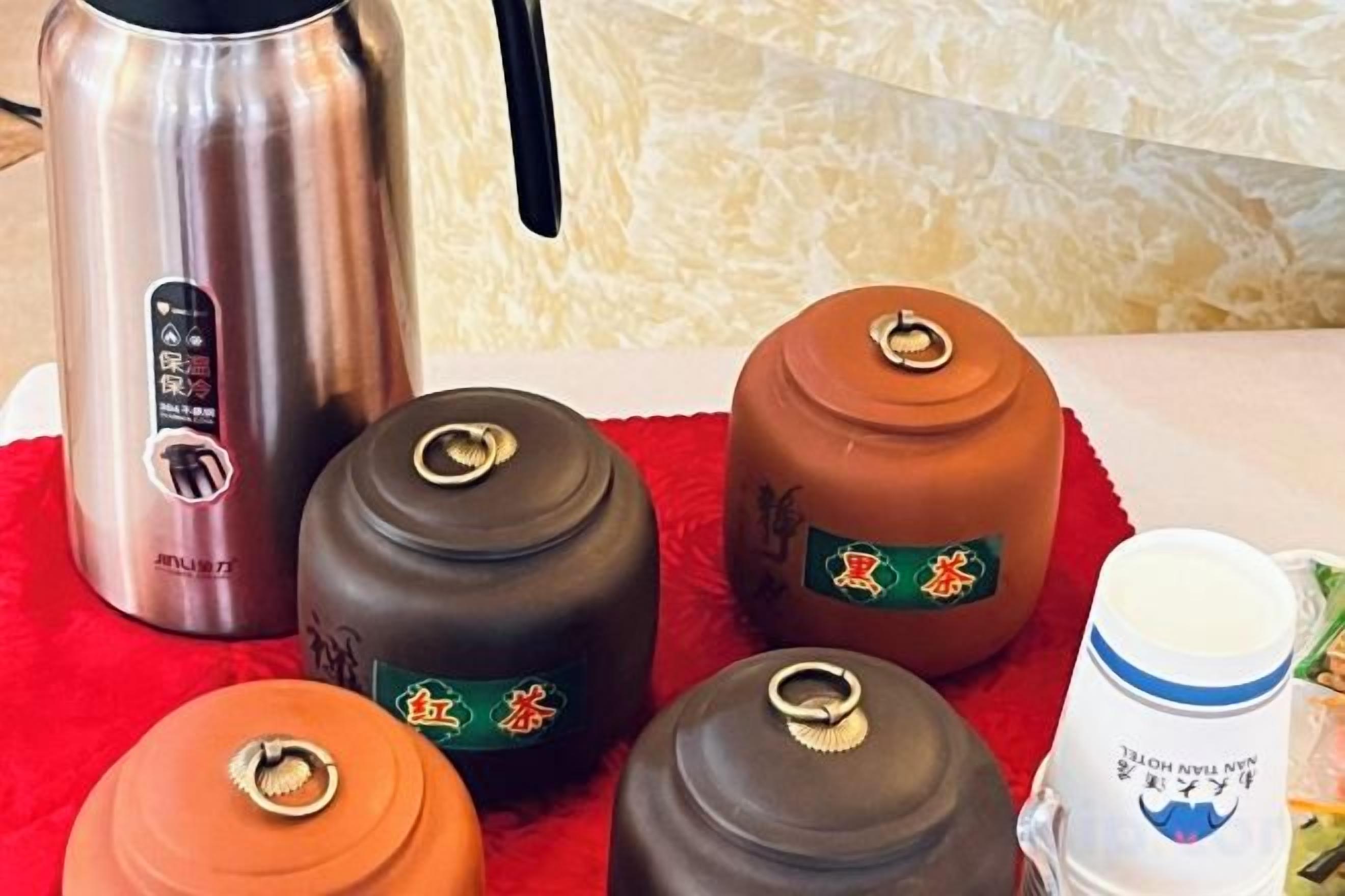 A stainless steel kettle, four ceramic containers with lids, and a white mug with a blue design on a red cloth.