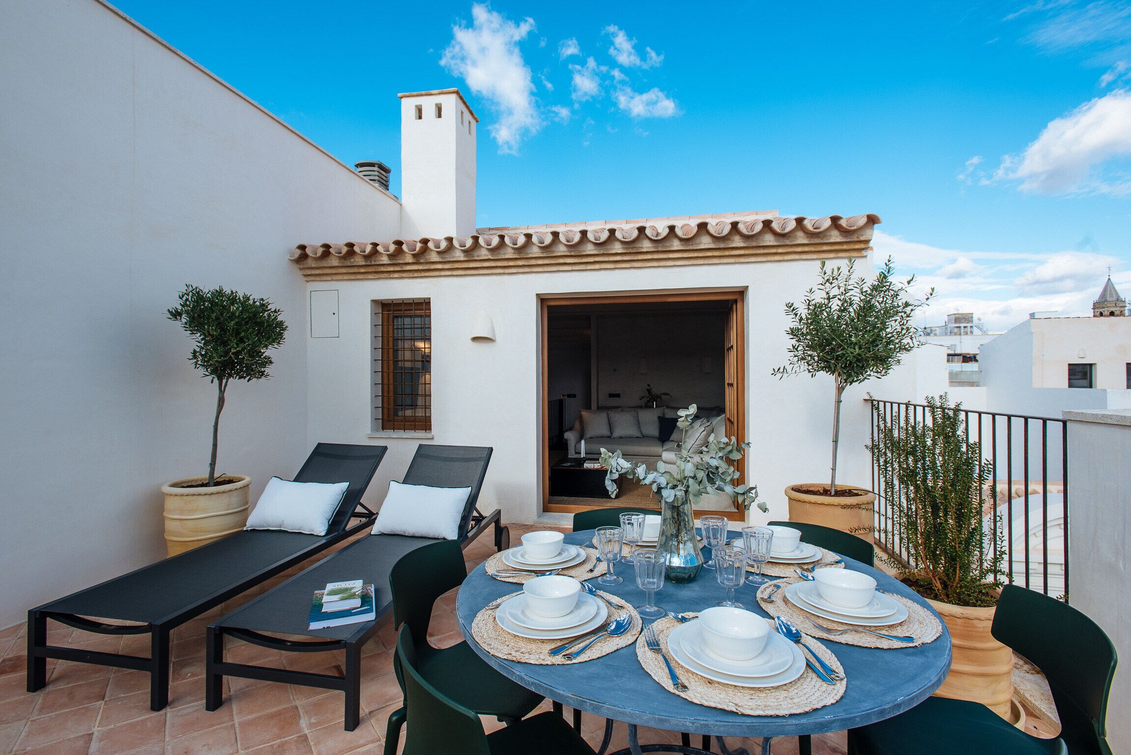 A rooftop terrace with a dining table set for six, surrounded by lounge chairs and potted plants.