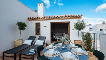 A rooftop terrace with a dining table set for six, surrounded by lounge chairs and potted plants.