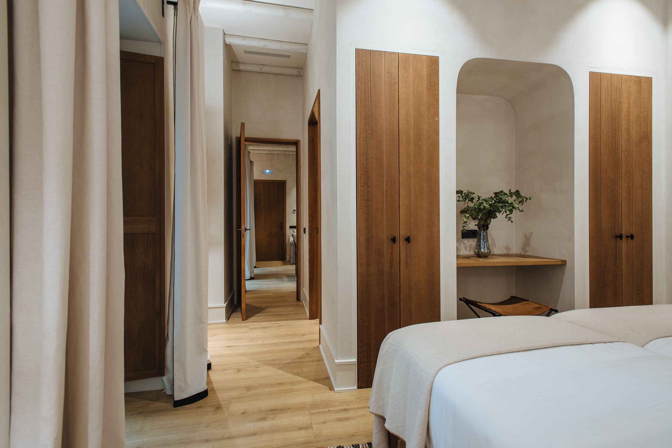 A hotel room with a wooden floor, a bed with white bedding, wooden wardrobes, and a small plant on a shelf.
