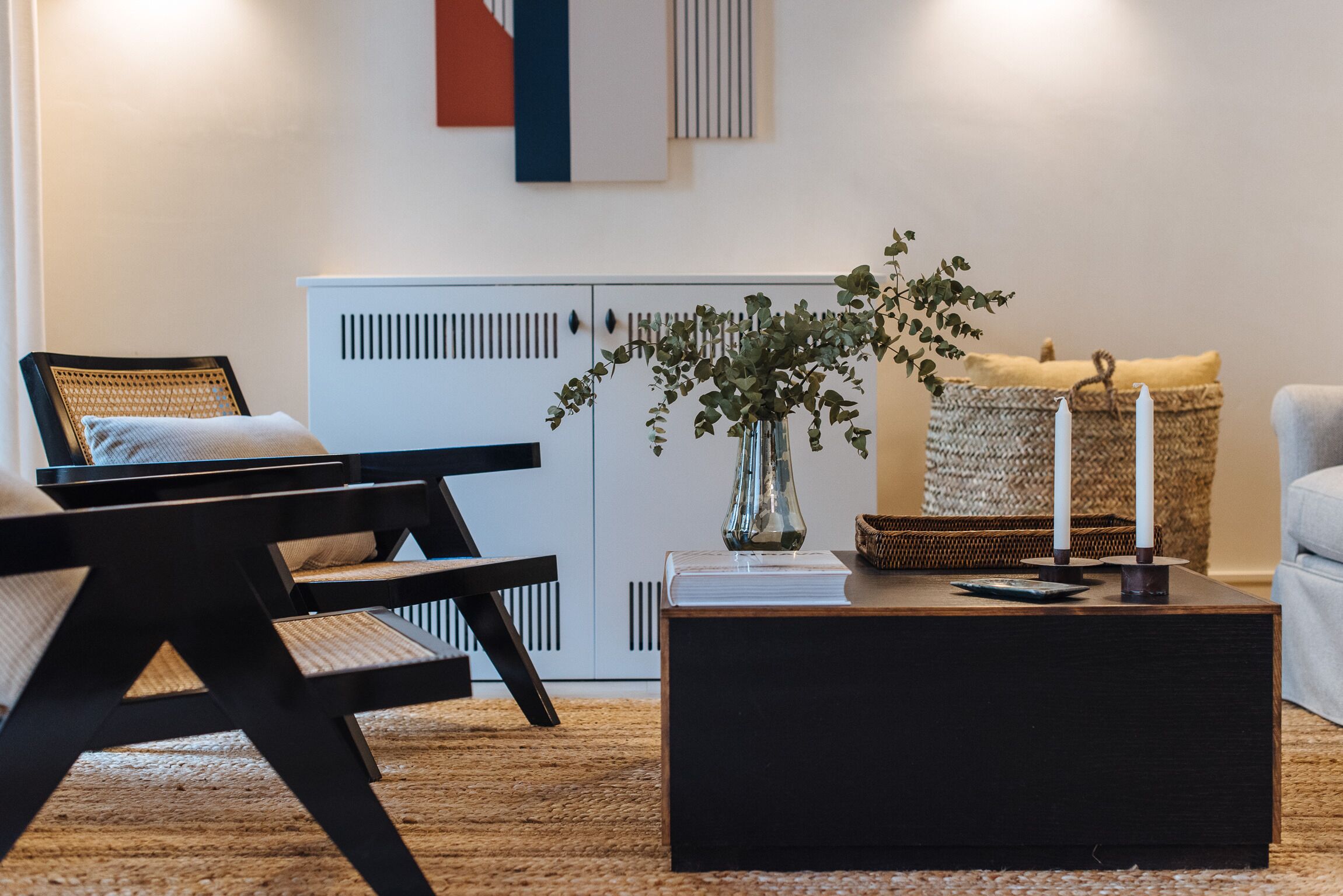 Una sala de estar moderna con un sofá negro, una mesa de centro negra, un sillón de mimbre, un armario blanco y un florero con follaje verde.