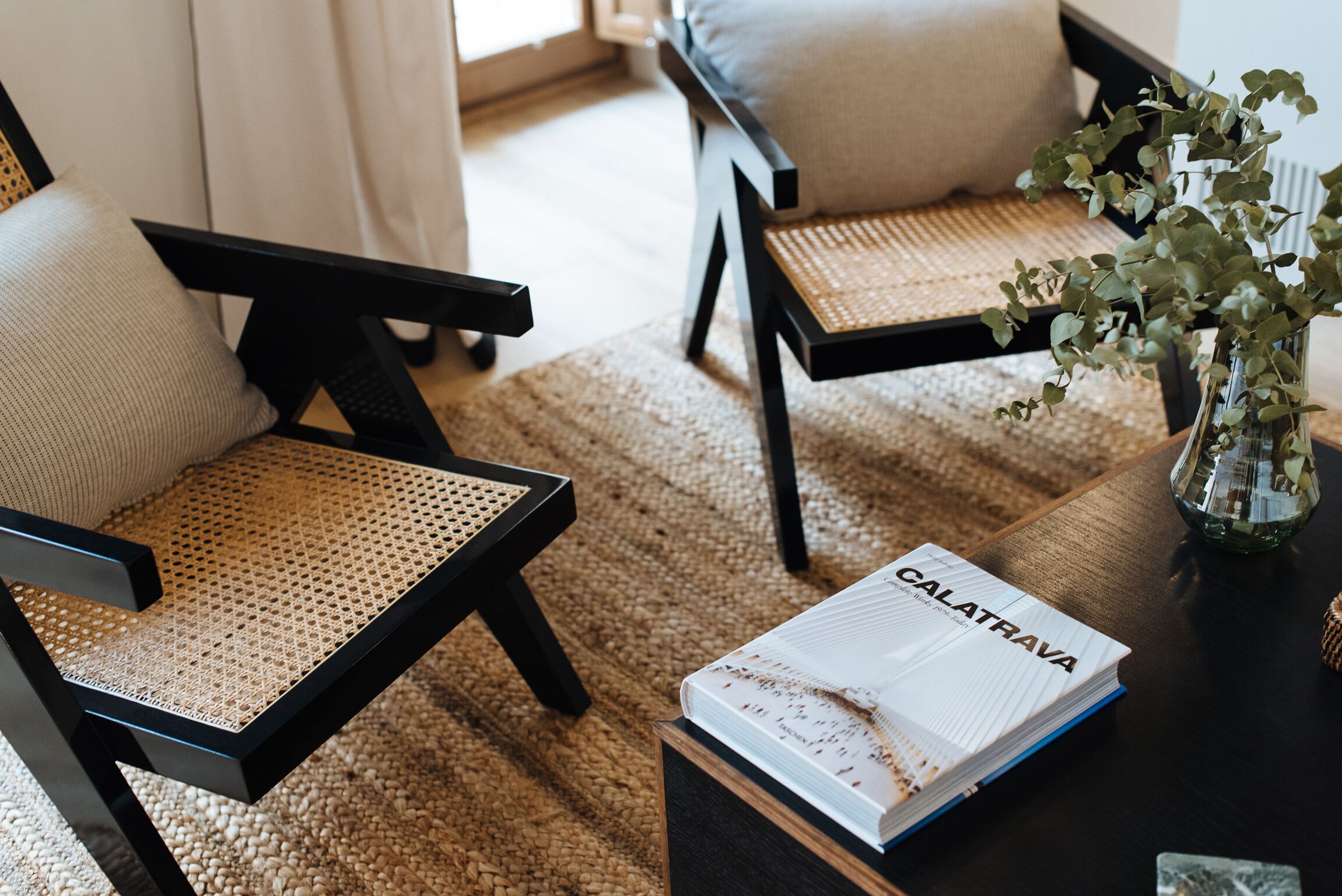 A room with two black chairs with woven seats, a small table with a book titled 'CALATRAVA', and a vase with green plants.