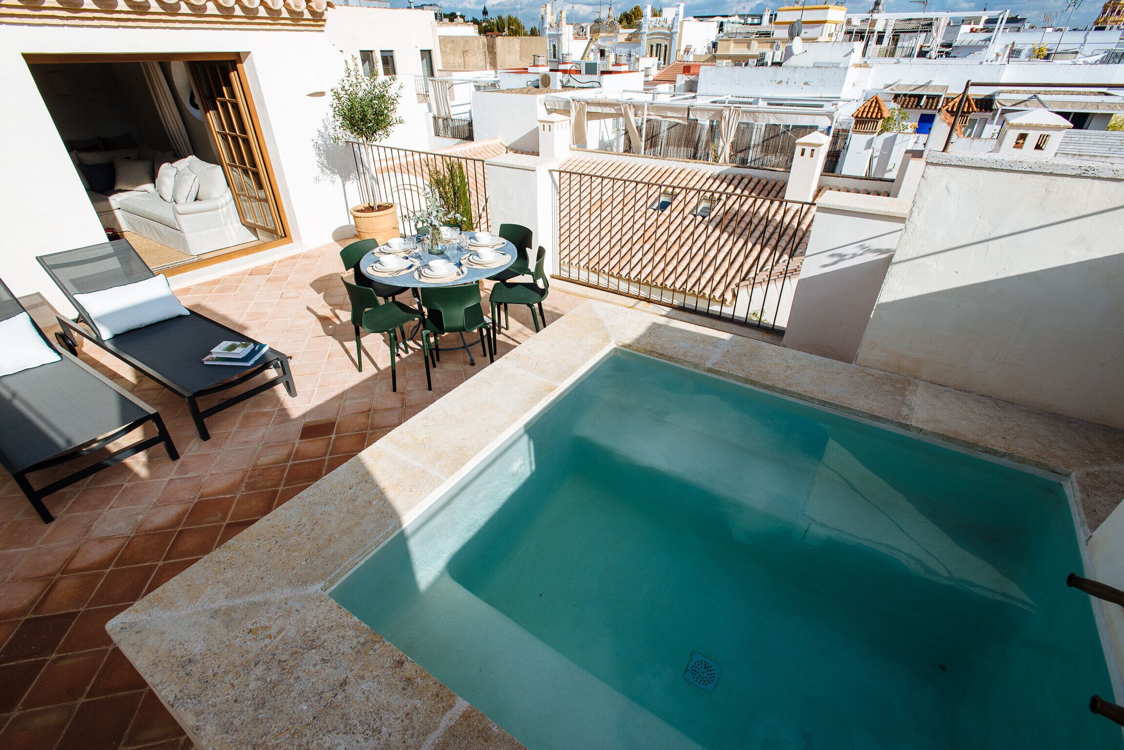 A rooftop pool area with a table and chairs, a lounge chair, and a view of the cityscape.