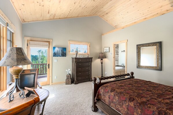 A bedroom with a wooden ceiling, a bed with a patterned bedspread, a dresser, a desk with a lamp, and a window with a view of trees.