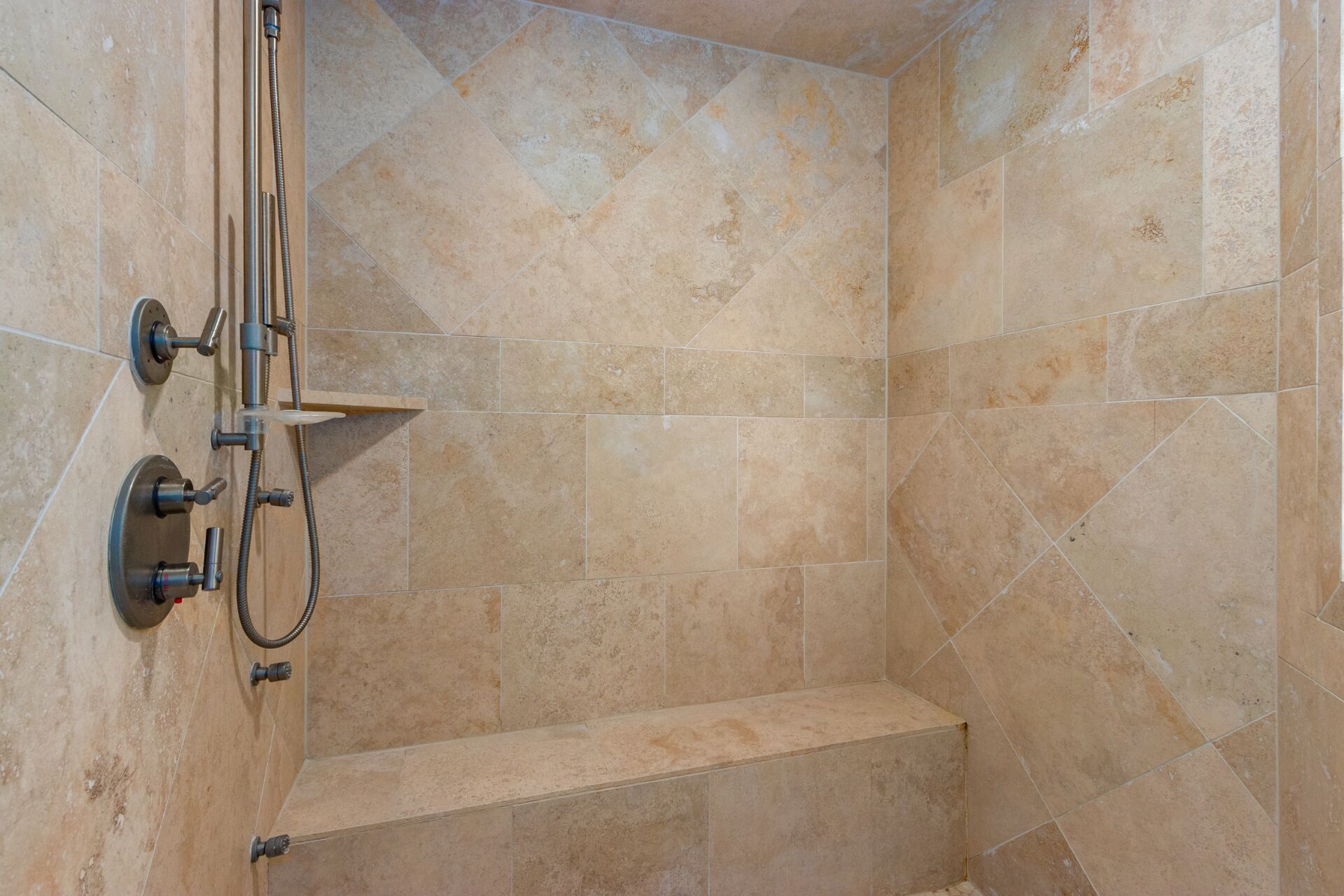 Zona de ducha con azulejos color beige, cabezal de ducha y un rociador de mano.